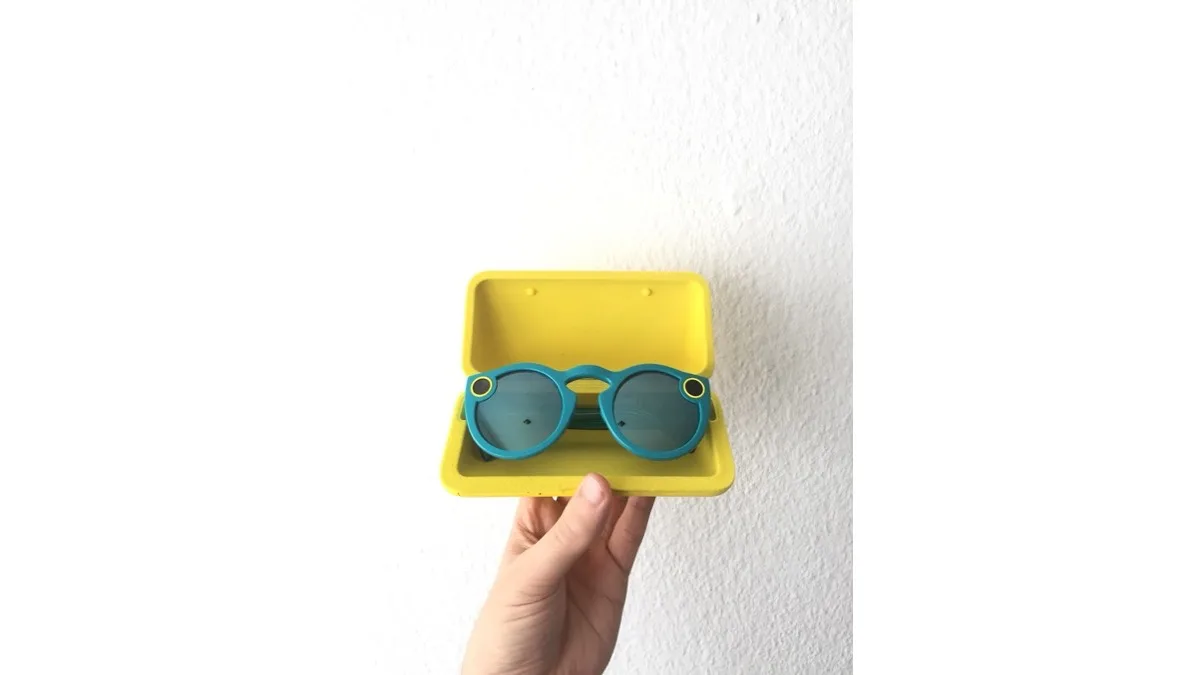 An earlier pair of teal Snap Spectacles sitting in their yellow charging case, held up against a white wall