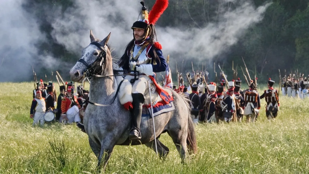 A cuirassier in a plumed helmet on a grey horse at a Battle of Waterloo reenactment, with lines of infantry reenactors and musket smoke behind him