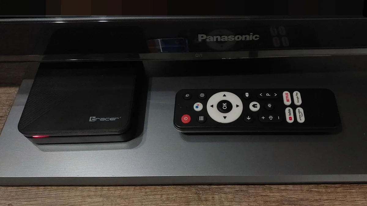 A black Android TV set-top box sits beside its remote on a stand in front of a television