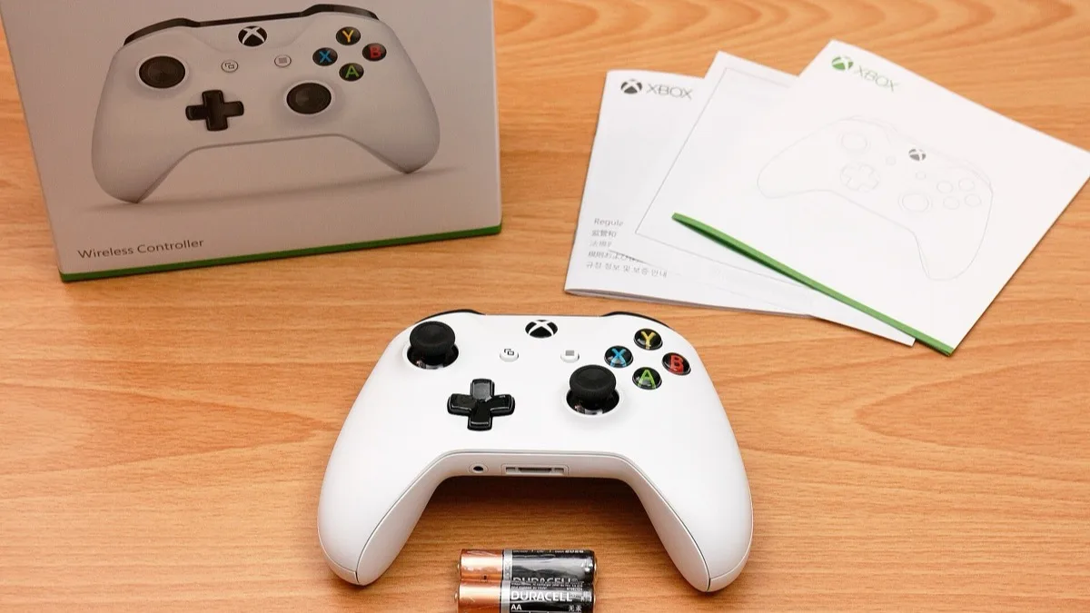 A white Xbox One wireless controller on a wooden desk beside its retail box, instruction leaflets and two AA batteries