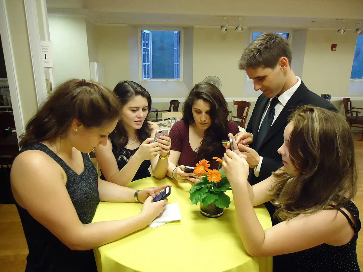 Five friends standing around a table, each texting on a smartphone