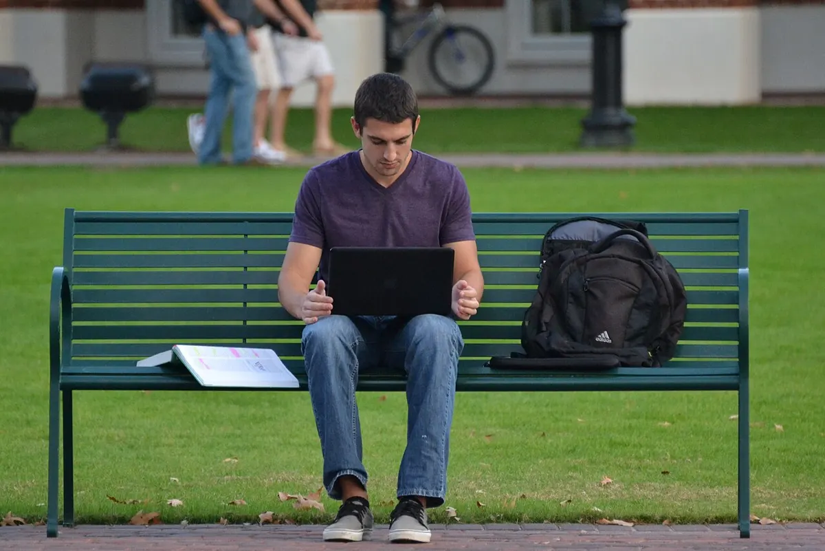 Student working on a laptop on a park bench with a backpack beside him