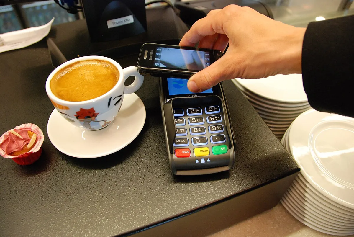 Hand holding a Samsung phone over a card payment terminal next to a cup of coffee